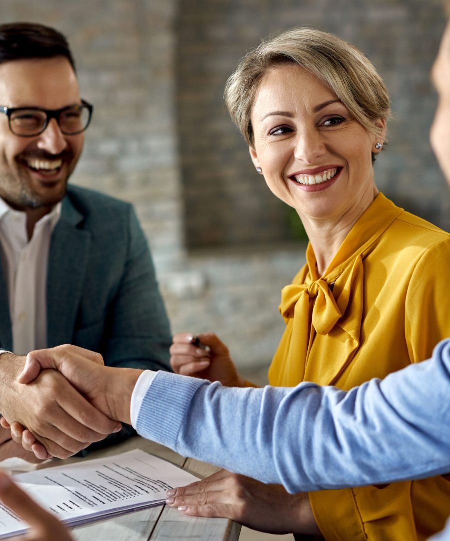 Happy woman signing a contract while her husband is handshaking with real estate agent in the office.